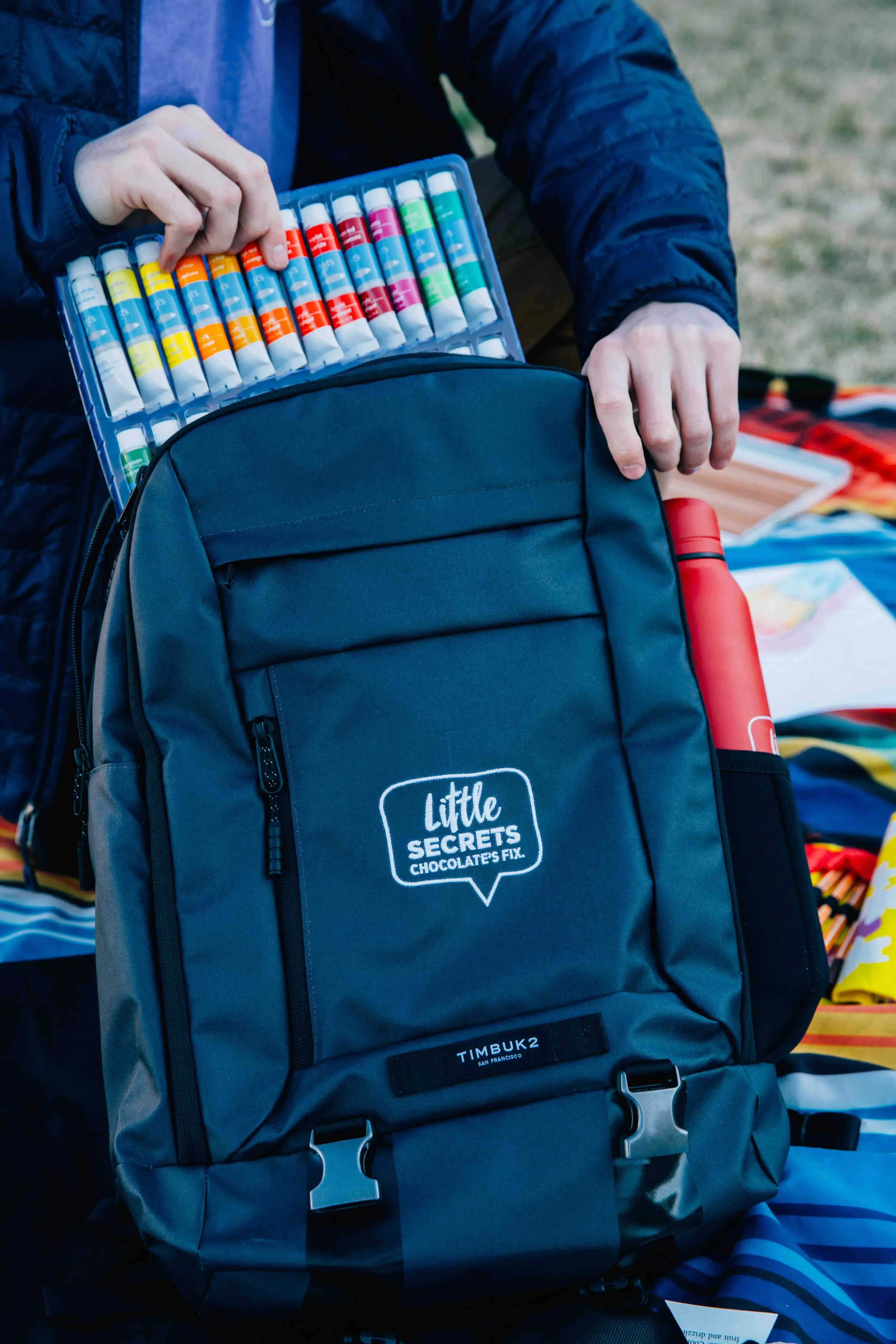 Person pull out paints from a custom Timbuk2 black backpack with embroidered Little Secrets logo